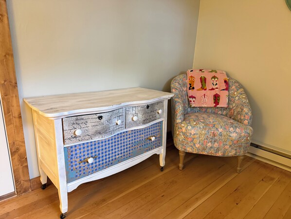 Une pièce avec un plancher en bois, une commode blanche aux tiroirs ornés de motifs bleus et un fauteuil à fleurs recouvert d’une couverture rose.