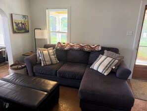 A living room with a dark-colored sofa, a black leather ottoman, and a painting of cows on the wall. - Wonderful 2-bedroom farmhouse in Salida.  Located @  Historic Hutchinson Ranch (Salida)
