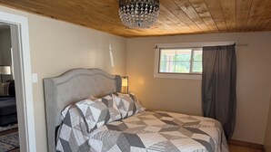A bedroom with a bed, a window with curtains, a chandelier, and a wooden ceiling.