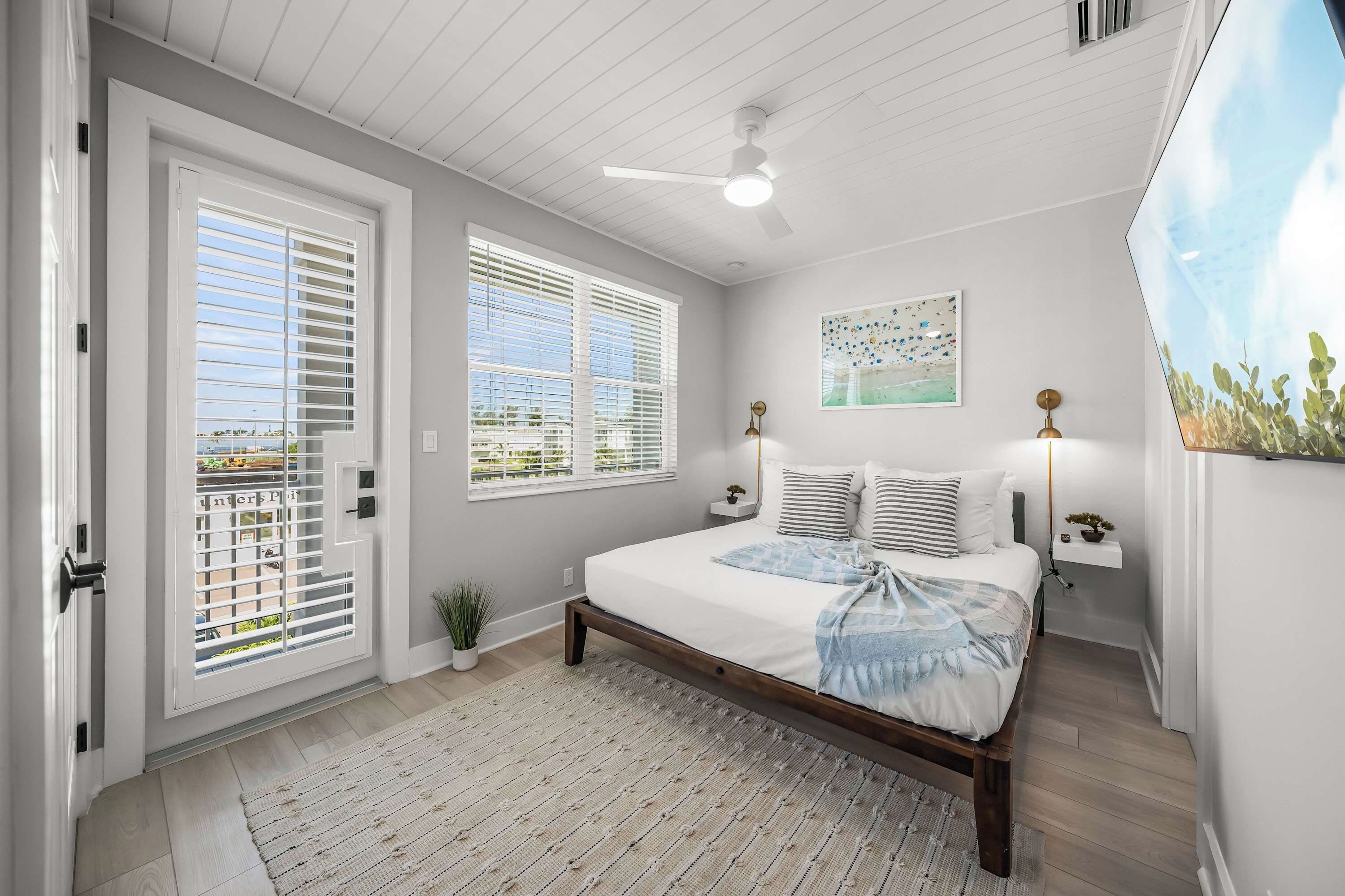 A bedroom with a bed, two windows, a ceiling fan, a painting, and a small plant.