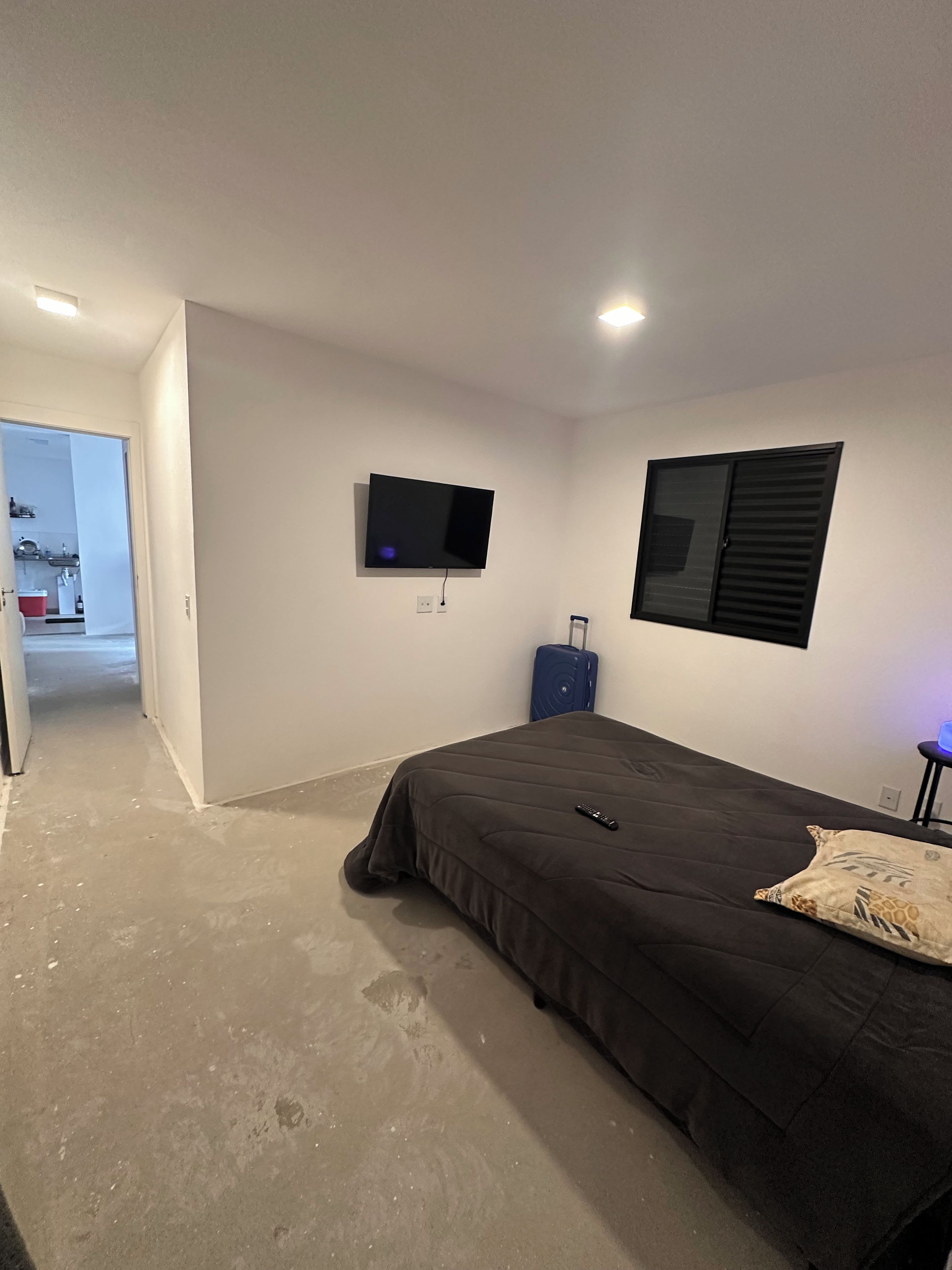 Ein Schlafzimmer mit einem Bett, einem an der Wand montierten Fernseher, einem Fenster mit Rollläden und einem Koffer.