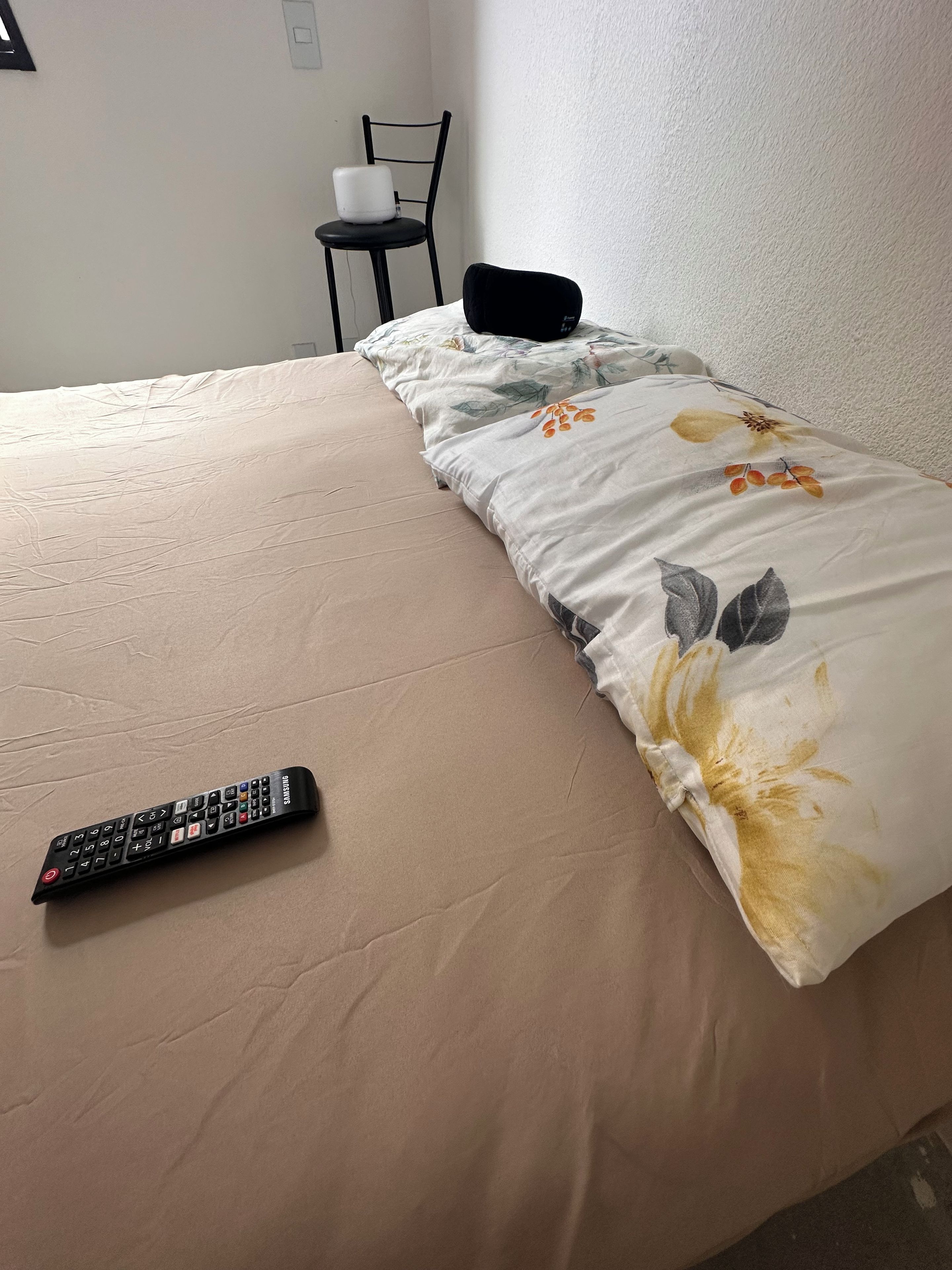 Un lit avec une couette à motifs floraux, une télécommande posée sur le lit et une chaise au fond de la pièce.