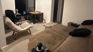 Uma sala de estar moderna com um sofá, uma poltrona, uma mesinha de centro com um copo e um controle remoto, e um espaço de jantar com mesa e cadeiras.