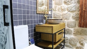 A bathroom with a white toilet, a wooden vanity with a sink, and a stone wall. - Katmani Hotel (ARTUKLU)
