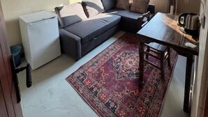 A compact living space with a sofa, a small table, a refrigerator, and a window with curtains. - Katmani Hotel (ARTUKLU)