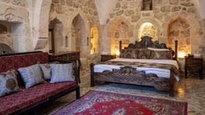 A room with a stone wall, a bed with an ornate headboard, a red sofa, and a patterned rug. - Katmani Hotel (ARTUKLU)