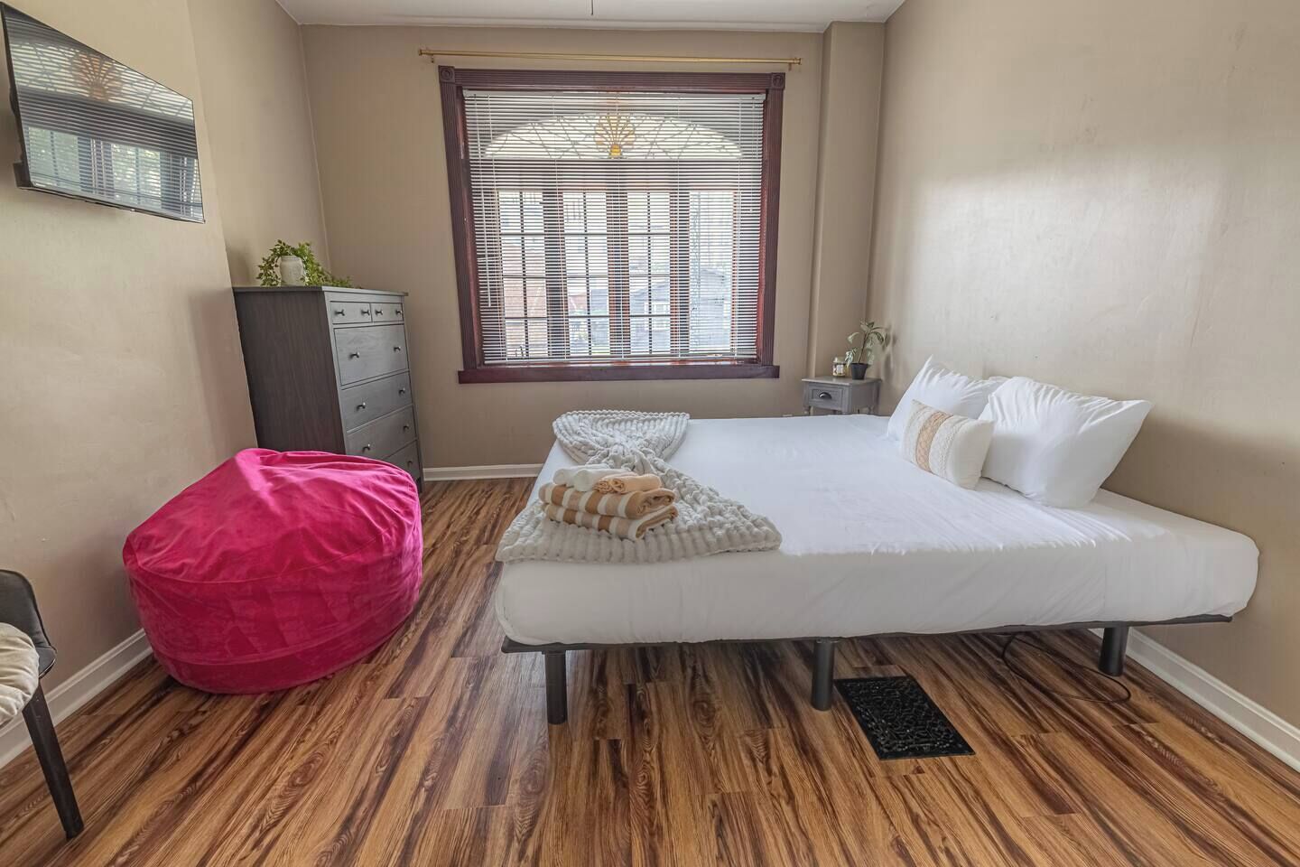 A bedroom with a bed, a red bean bag chair, a dresser, a window with blinds, and a small nightstand.