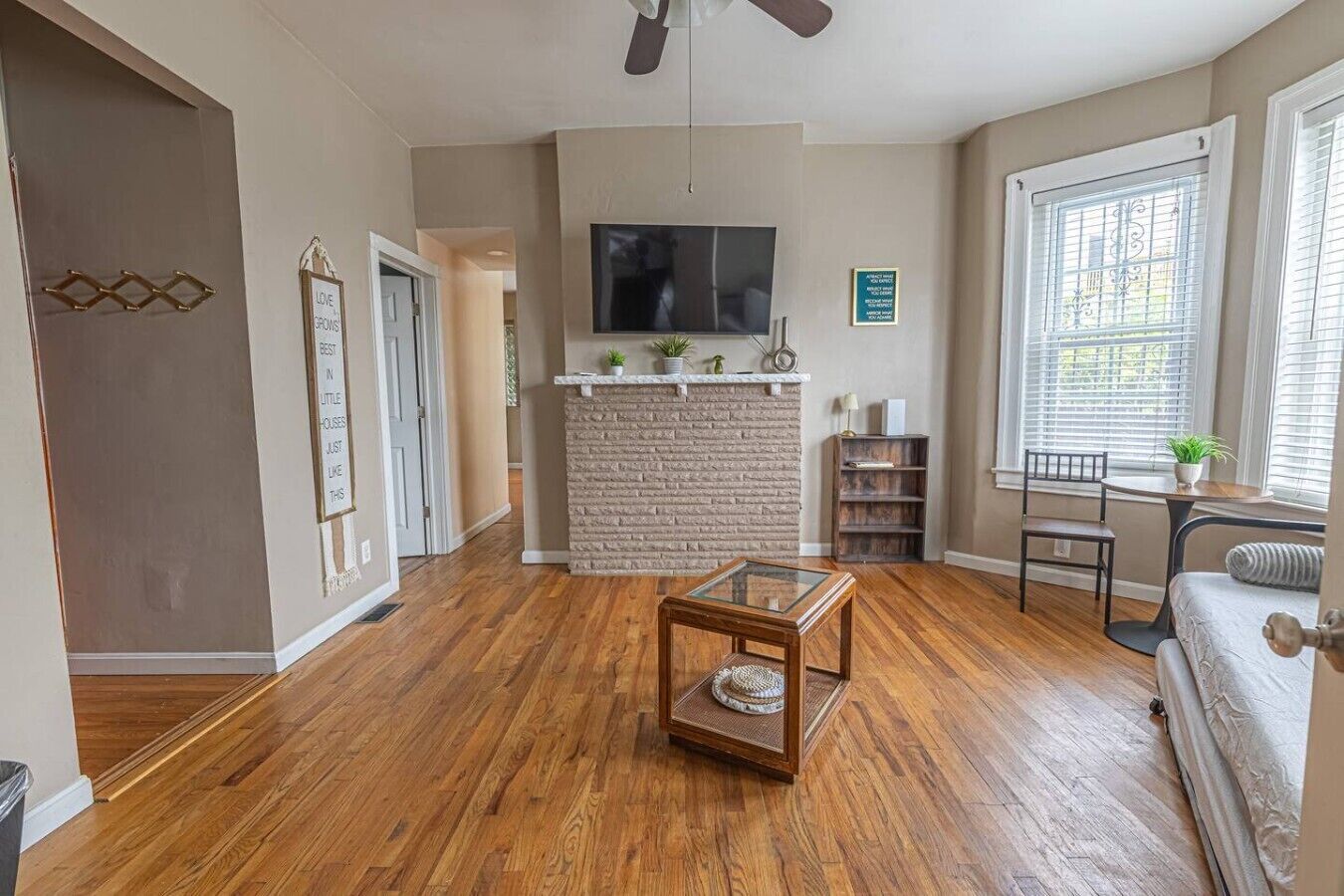 A living room with hardwood flooring, a stone fireplace, a flat-screen TV, and a window with blinds.