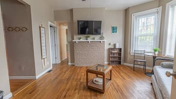 A living room with hardwood flooring, a stone fireplace, a flat-screen TV, and a window with blinds.