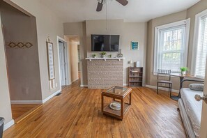 A living room with hardwood flooring, a stone fireplace, a flat-screen TV, and a window with blinds. - Cozy Mount Washington 1BR with Private Patio (Pittsburgh)