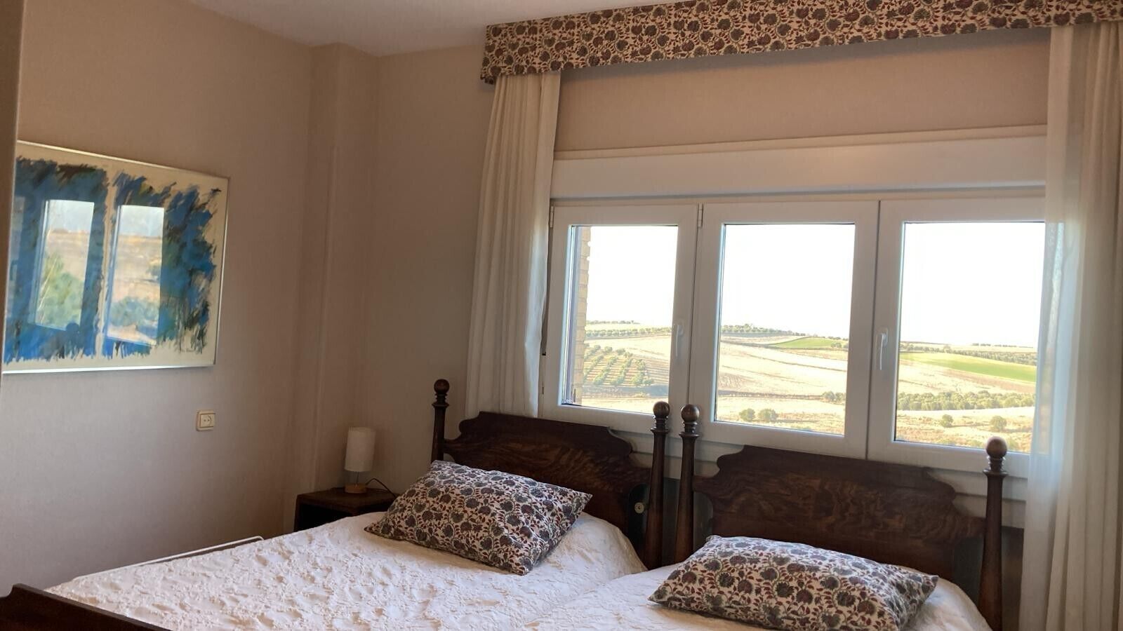 Een slaapkamer met een groot bed, twee kussens, een nachtkastje, een lamp en een raam met uitzicht op velden.