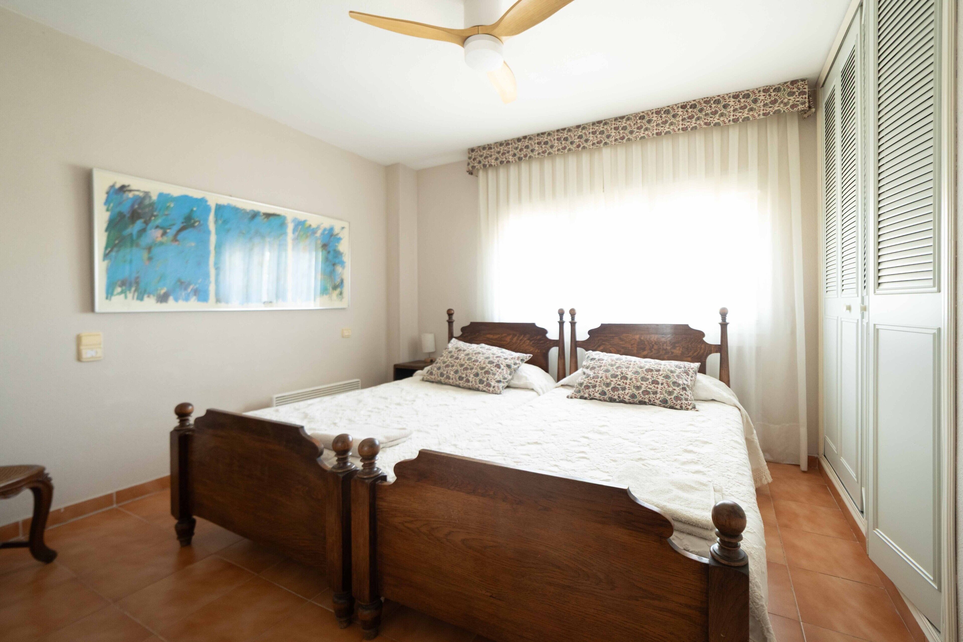 Une chambre avec un lit en bois, un ventilateur de plafond, une grande fenêtre avec des rideaux et un tableau accroché au mur.