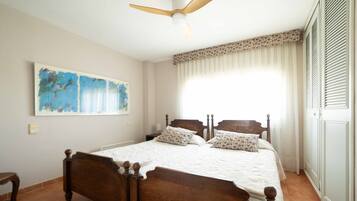 Une chambre avec un lit en bois, un ventilateur de plafond, une grande fenêtre avec des rideaux et un tableau accroché au mur.