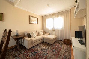 Un salon avec un canapé beige, un tapis à motifs, un téléviseur et un tableau encadré accroché au mur.
