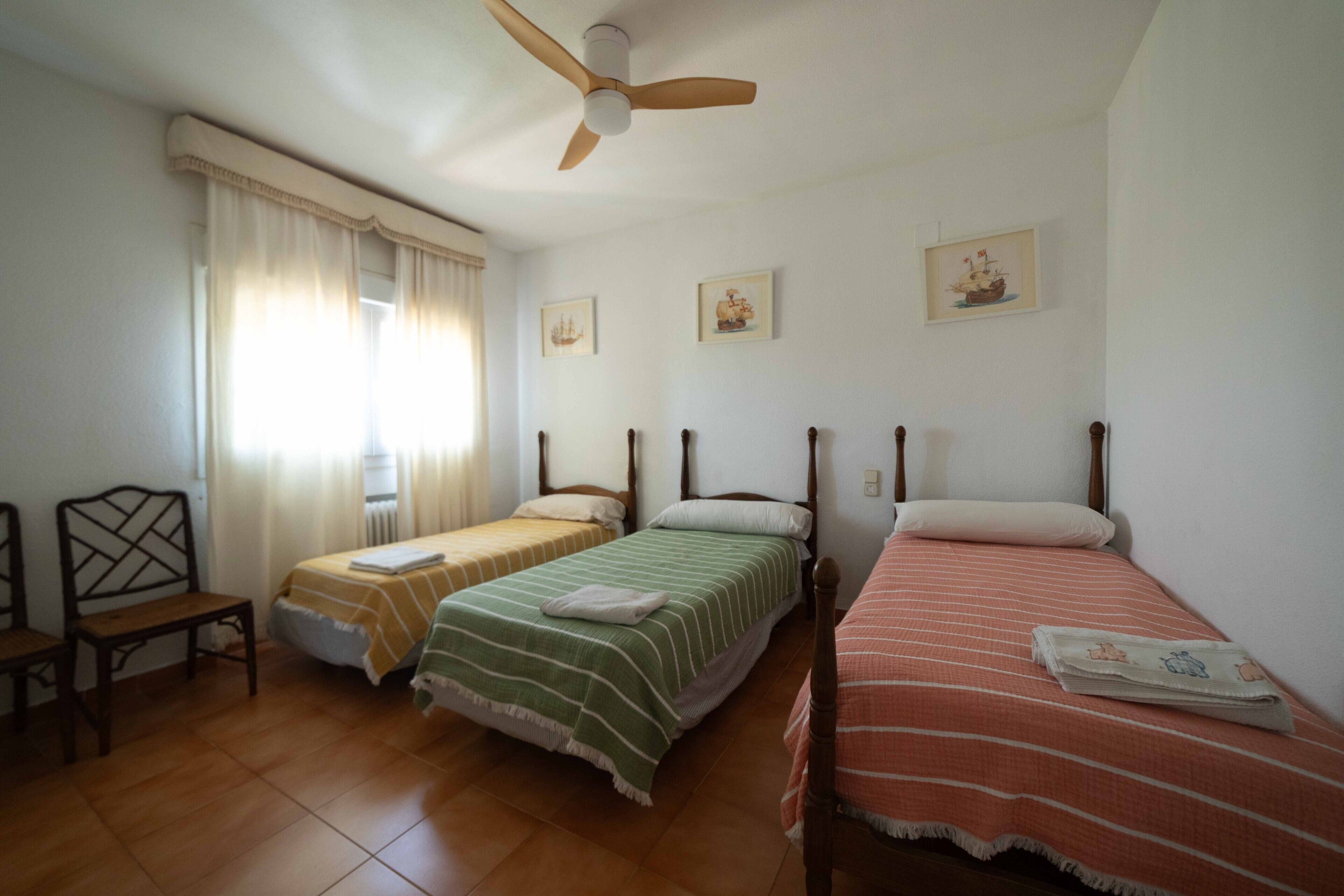 Een kamer met drie eenpersoonsbedden, een plafondventilator, een raam met gordijnen en een stoel.
