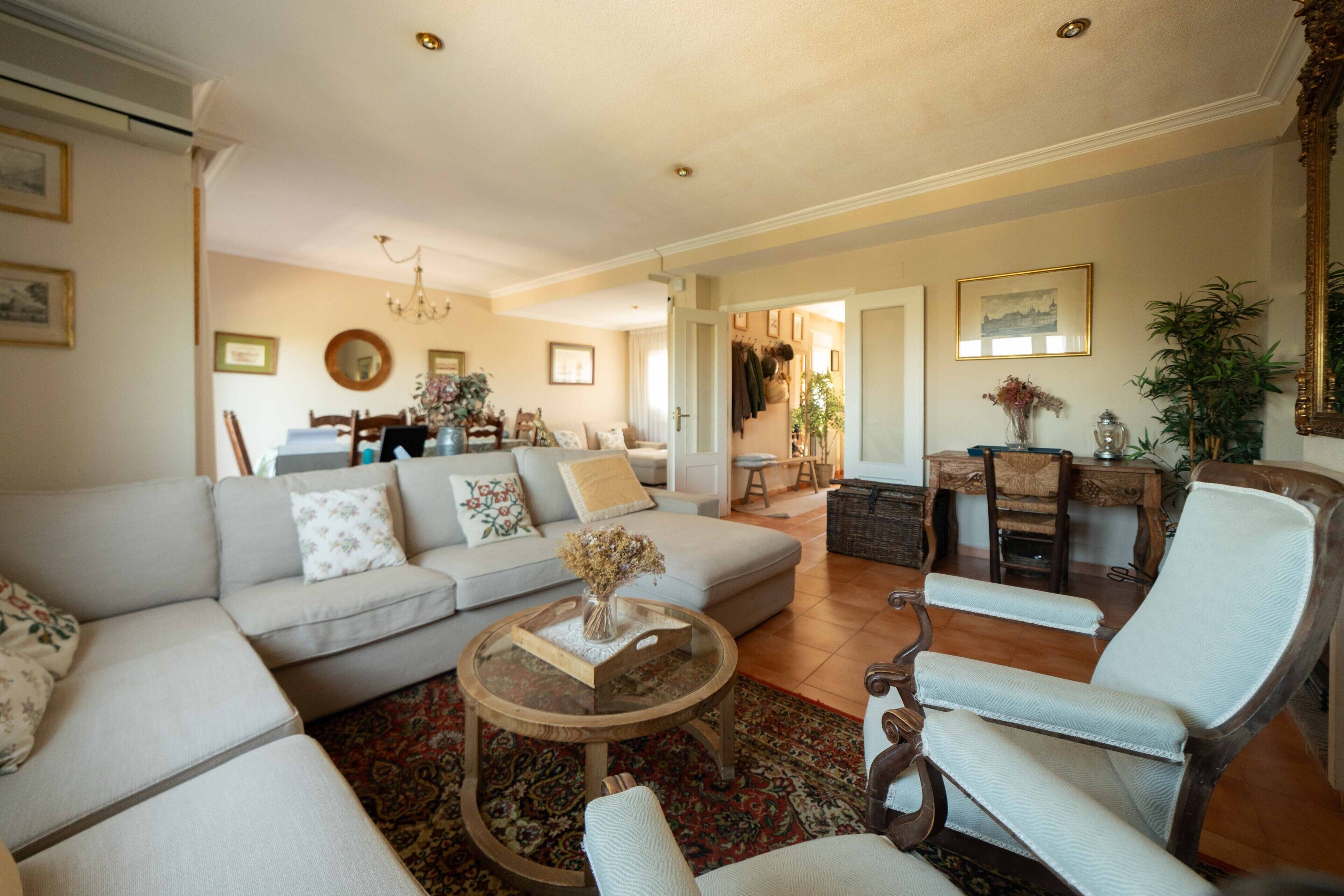 A spacious living room with a large sofa, a coffee table, and a chair. There is a dining area in the background with a chandelier.