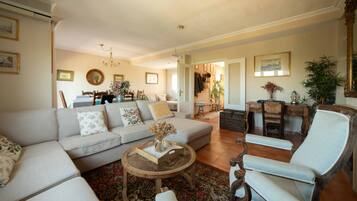 A spacious living room with a large sofa, a coffee table, and a chair. There is a dining area in the background with a chandelier.
