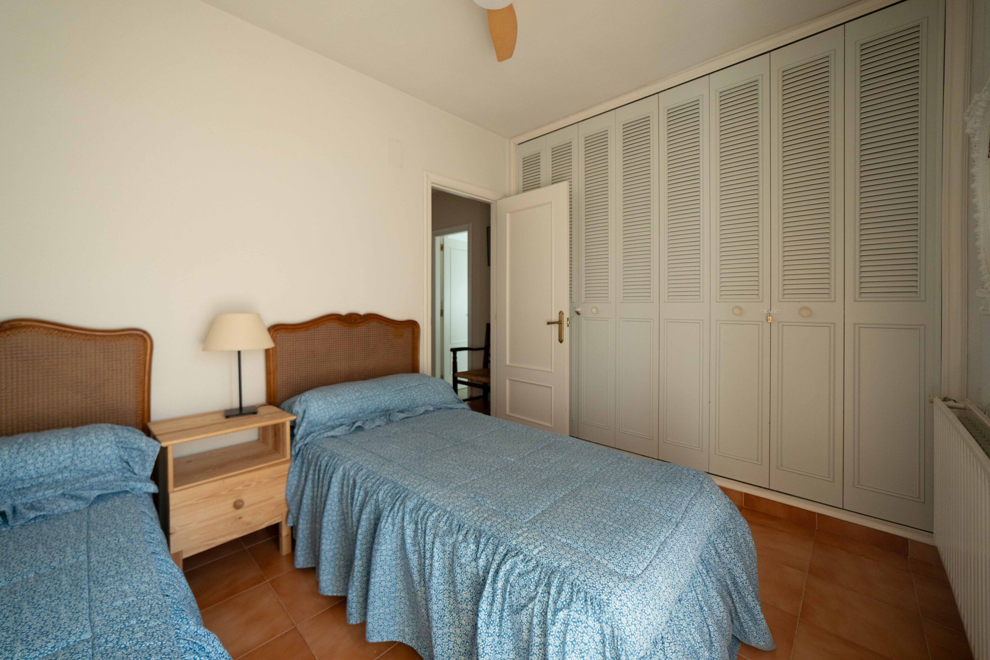 Un dormitorio con dos camas individuales, una mesita de noche con lámpara y un armario con puertas batientes.
