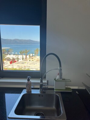 A kitchen with a stainless steel sink, a window with a view of a beach and mountains, and a bottle of hand soap. - Sunset Resort Uji Ftohtë, Vlorë (Uji Ftohte, Vlorë)