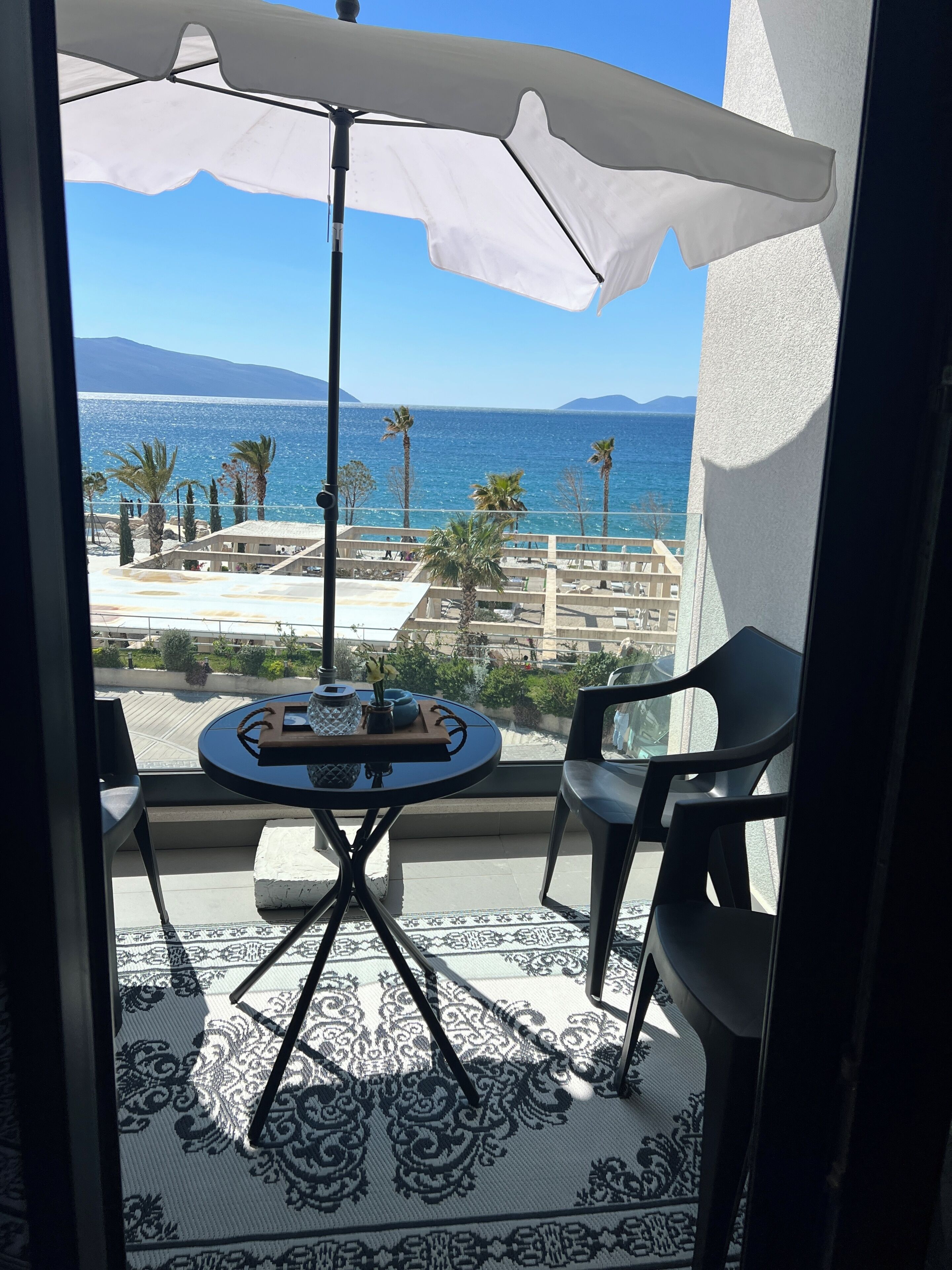 A balcony with a table, chairs, and an umbrella overlooking a coastal view with palm trees and a distant mountain.