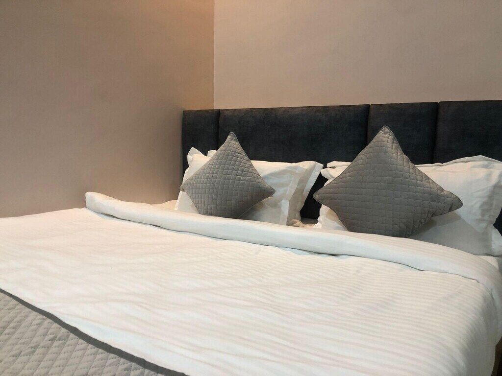Un letto rifatto con copriletto bianco e cuscini grigi, una testata scura e un muro neutro sullo sfondo.