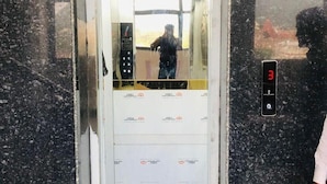 Un ascensor con una pantalla digital que muestra el número 3, una persona parada afuera y el reflejo de un edificio en la puerta del ascensor.