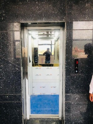 An elevator with a digital display showing the number 3, a person standing outside, and a reflection of a building in the elevator door.