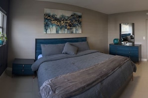 A bedroom with a bed, a dresser, a nightstand, a mirror, and a wall-mounted abstract painting. - 2BR/2BA by Convention Ctr, Walk to Beach + Parking (Miami Beach)