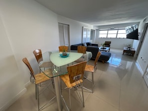 A modern living room with a glass dining table surrounded by wooden chairs, a sofa, and a TV mounted on the wall. - 2BR/2BA by Convention Ctr, Walk to Beach + Parking (Miami Beach)