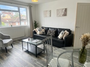 A modern living room with a black leather sofa, a glass coffee table, a grey armchair, and a dining table with chairs. There are two abstract paintings on the wall and a view of the outside through a large window. - Wimbledon 2br Free Parking Fast Wi-fi Near Tube (London)