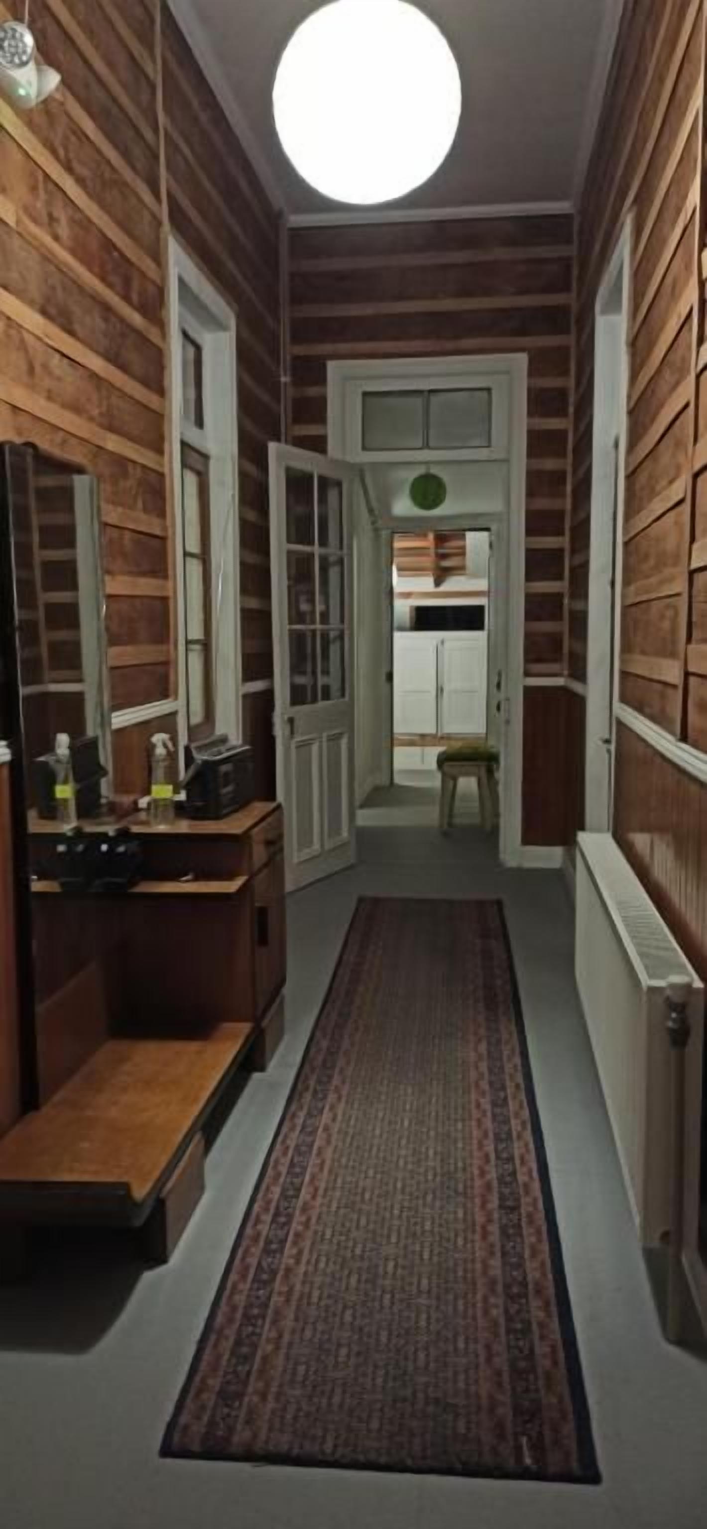 A narrow hallway with wooden walls, a rug, a mirror, and a door leading to another room.