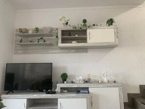 A living room with a flat-screen TV, a white cabinet, and shelves with decorative items and plants. - Charming apartment with views of the sea and mountains (Roses)