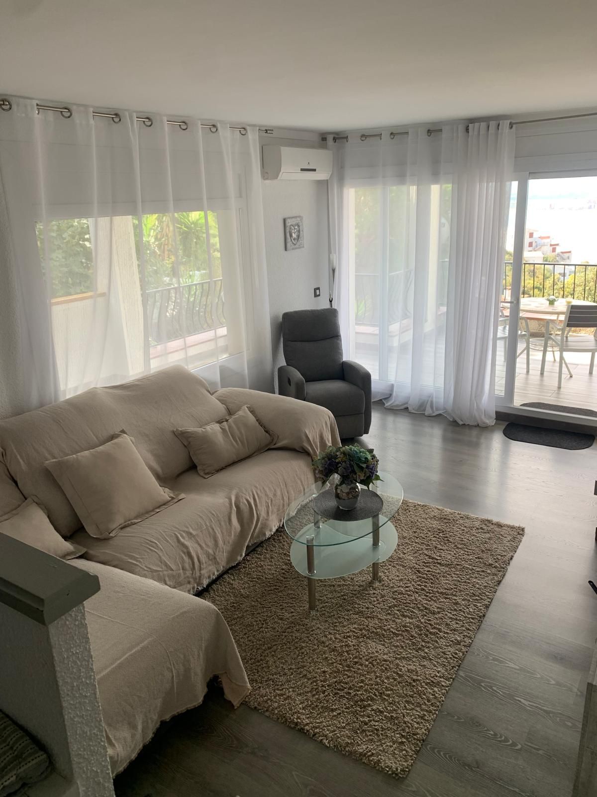Un salon comprenant un canapé, une table basse en verre avec un vase, et un fauteuil. On aperçoit un balcon avec une table et des chaises à travers les fenêtres.