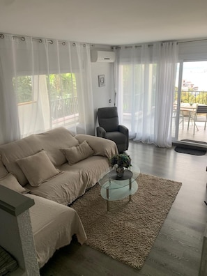 A living room with a sofa, a glass coffee table with a vase, and a chair. There is a balcony with a table and chairs visible through the windows. - Charming apartment with views of the sea and mountains (Roses)