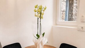 Mesa de madeira redonda com duas cadeiras estofadas pretas, um vaso branco com orquídeas verdes e uma parede de pedra visível através de uma janela.
