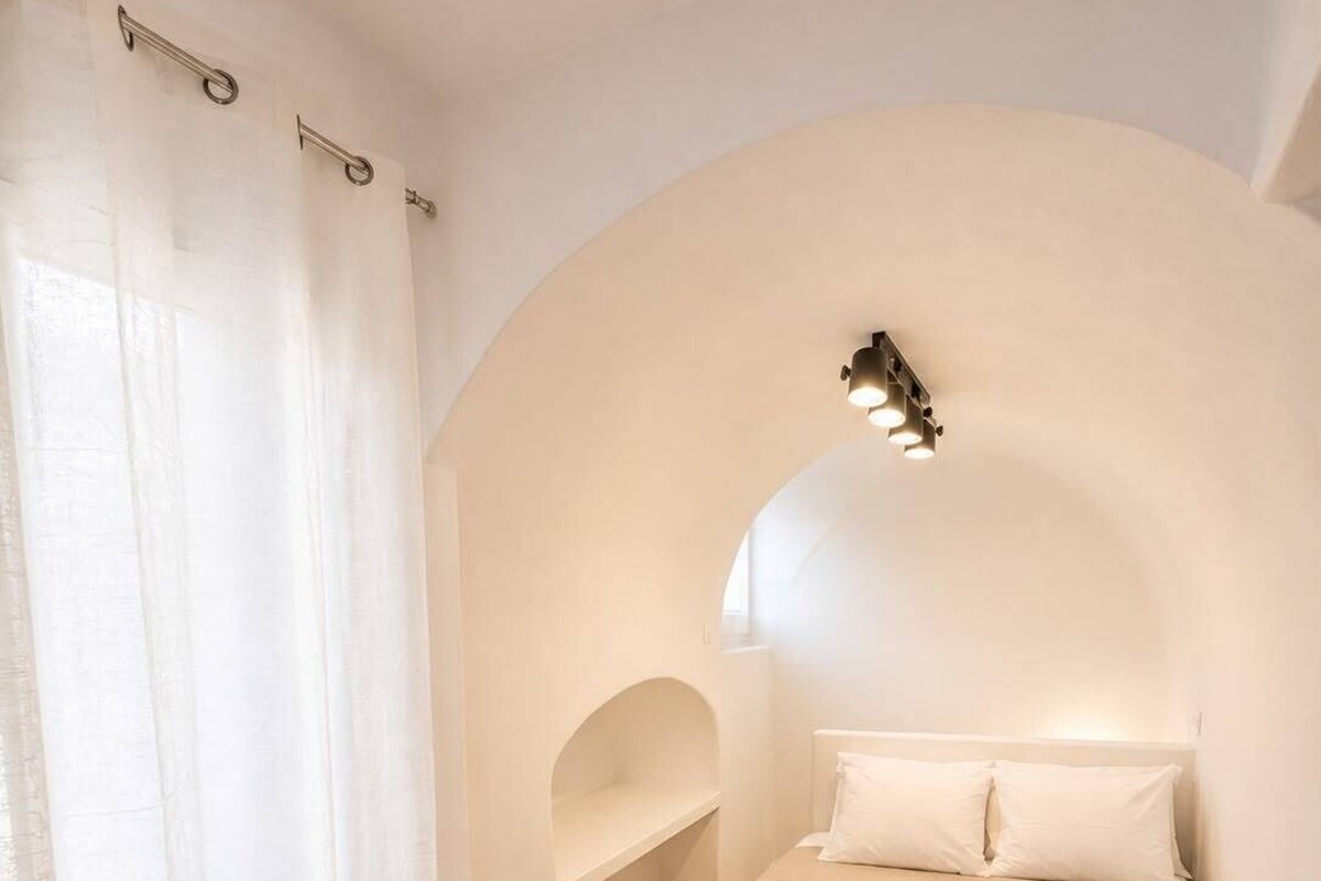 Un dormitorio con techo curvo, cama con colcha a cuadros, mesita de noche y ventana con cortinas translúcidas.