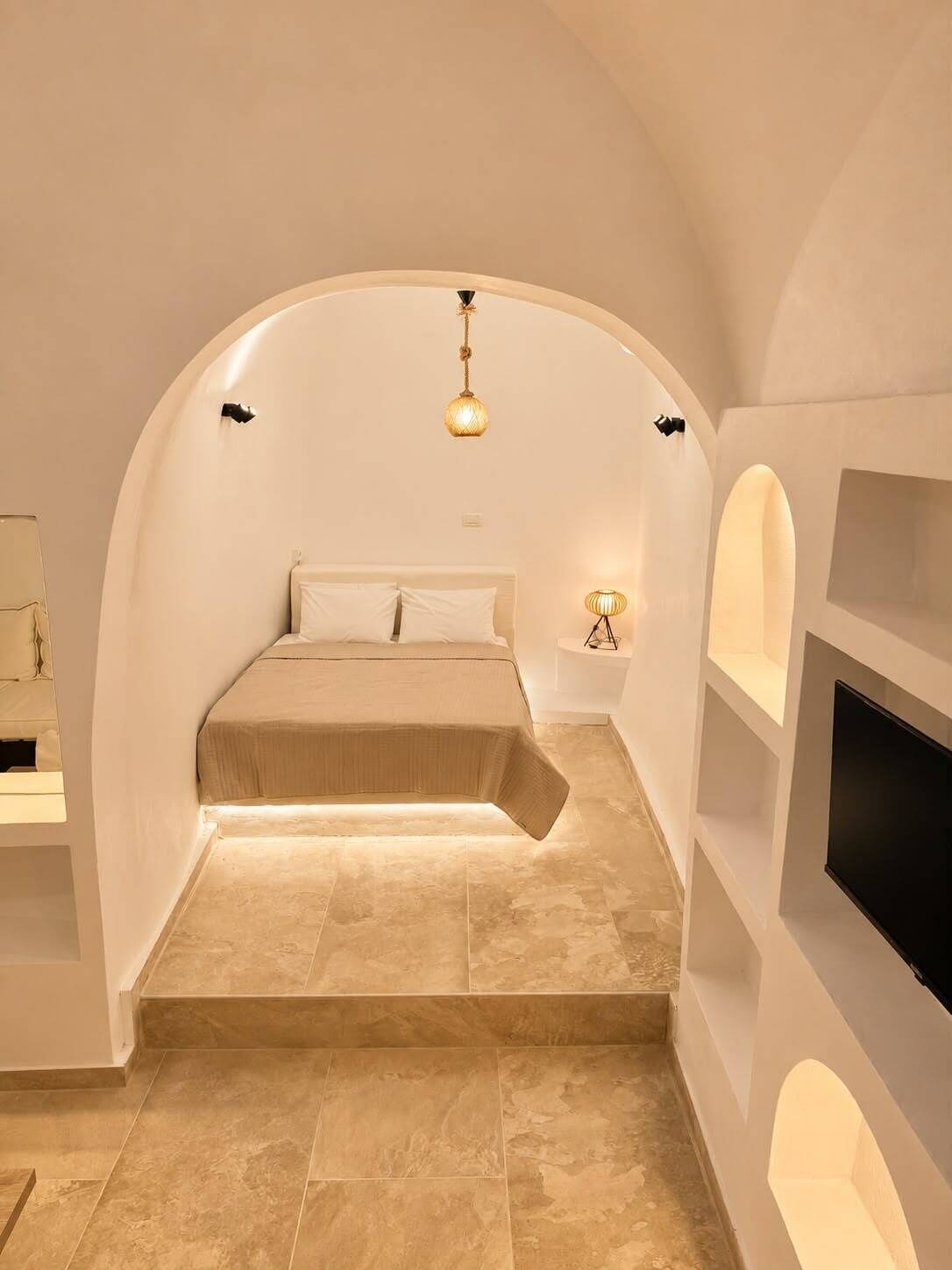 A bedroom with a curved ceiling, a bed with a beige cover, a wall-mounted TV, and a small bedside table with a lamp.