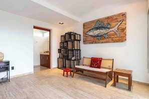 Ein Wohnzimmer mit einer hölzernen Bank, einem roten Hocker, einem Bücherregal und einem an der Wand befestigten Kunstwerk mit einem Fisch.