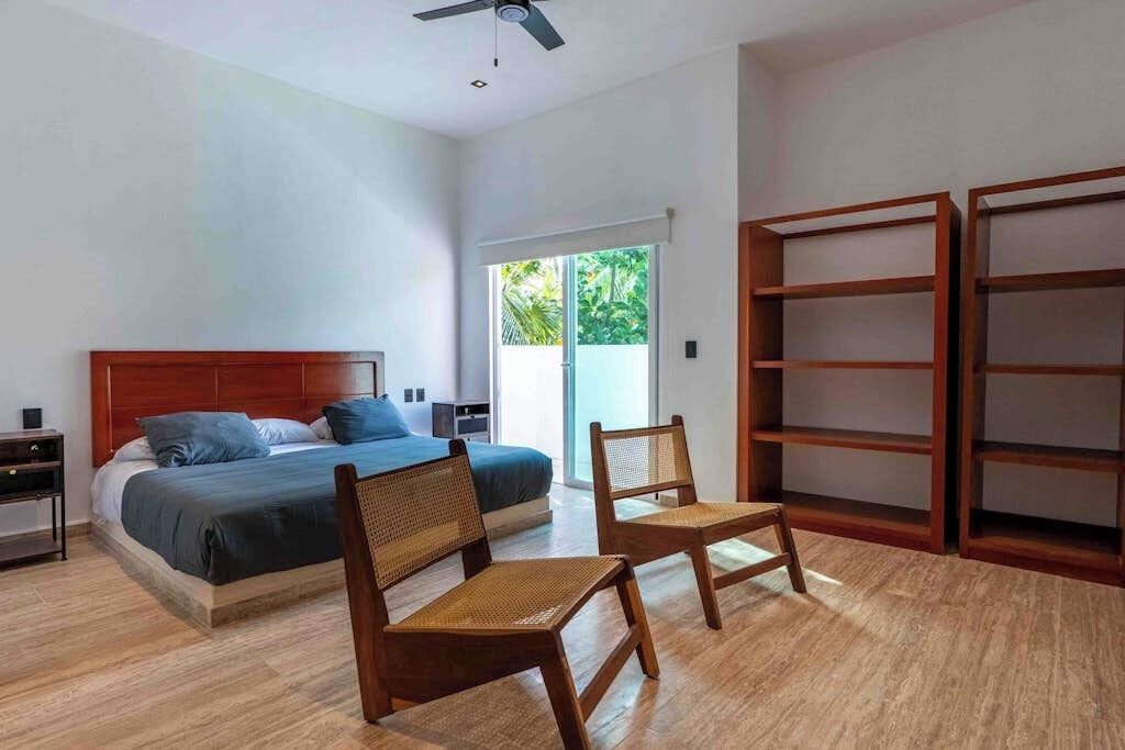 A bedroom with a bed, two chairs, a nightstand, a wardrobe, and a window with a view of greenery.