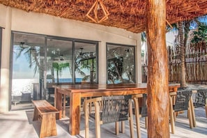 An outdoor seating area with a wooden table and chairs, large windows, and a view of the beach. - Beachfront home pool, chef, transport (Tulum)
