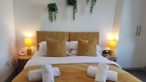 Quarto de hotel com cama, duas mesas de cabeceira, luminária, relógio e planta fixada na parede.