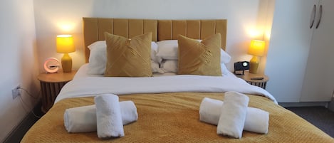 A hotel room with a bed, two bedside tables, a nightstand, a lamp, a clock, and a wall-mounted plant.