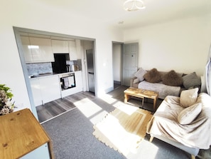 Een moderne keuken met witte kasten en een ingebouwde oven, een woonruimte met een bank en een salontafel, en een deur naar een andere kamer.
