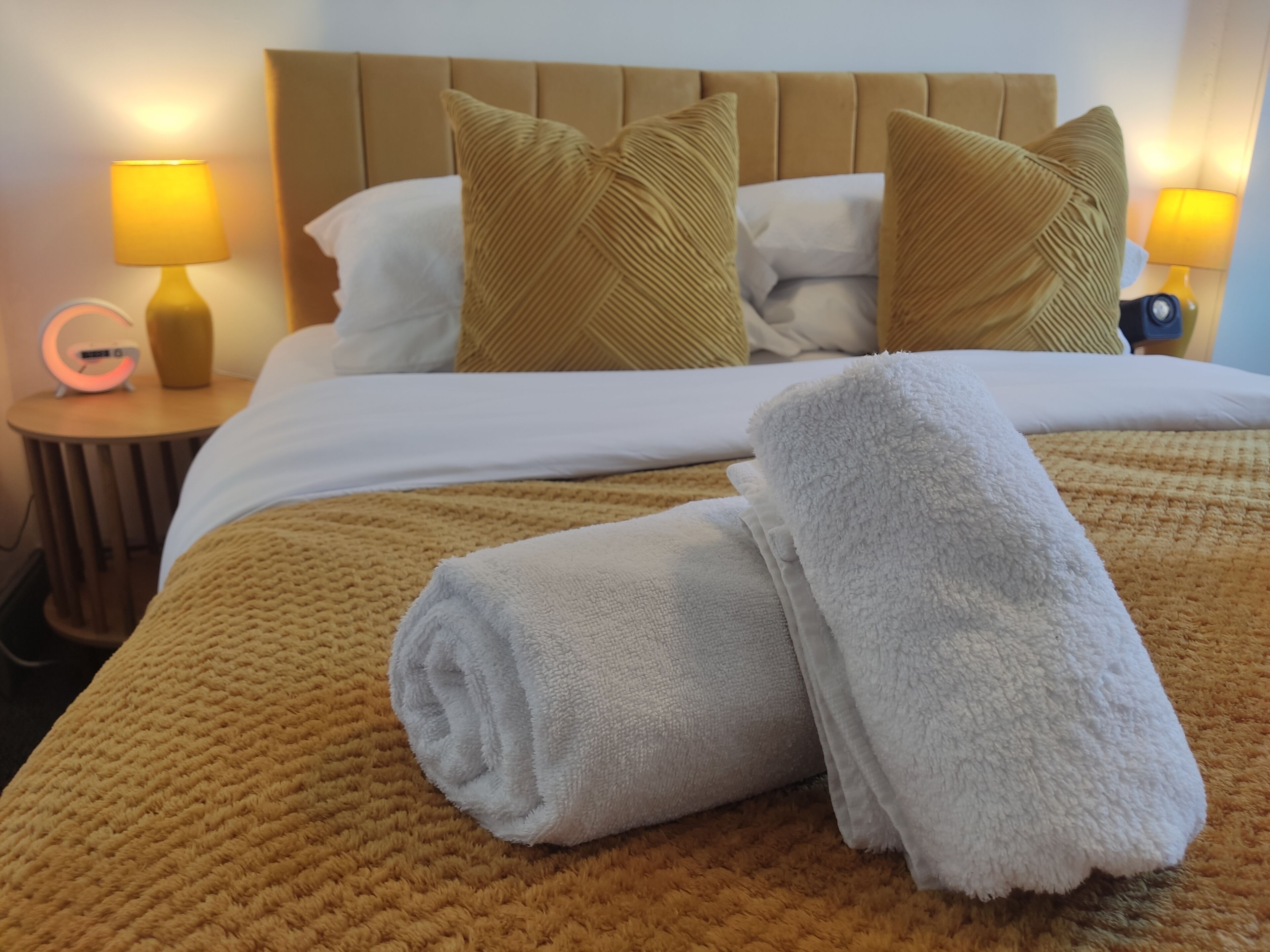 Een netjes opgemaakt bed met een bruine deken, twee kussens en een opgerolde witte handdoek. Twee bedlampen en een klein tafeltje met een lamp en een klok staan ernaast.