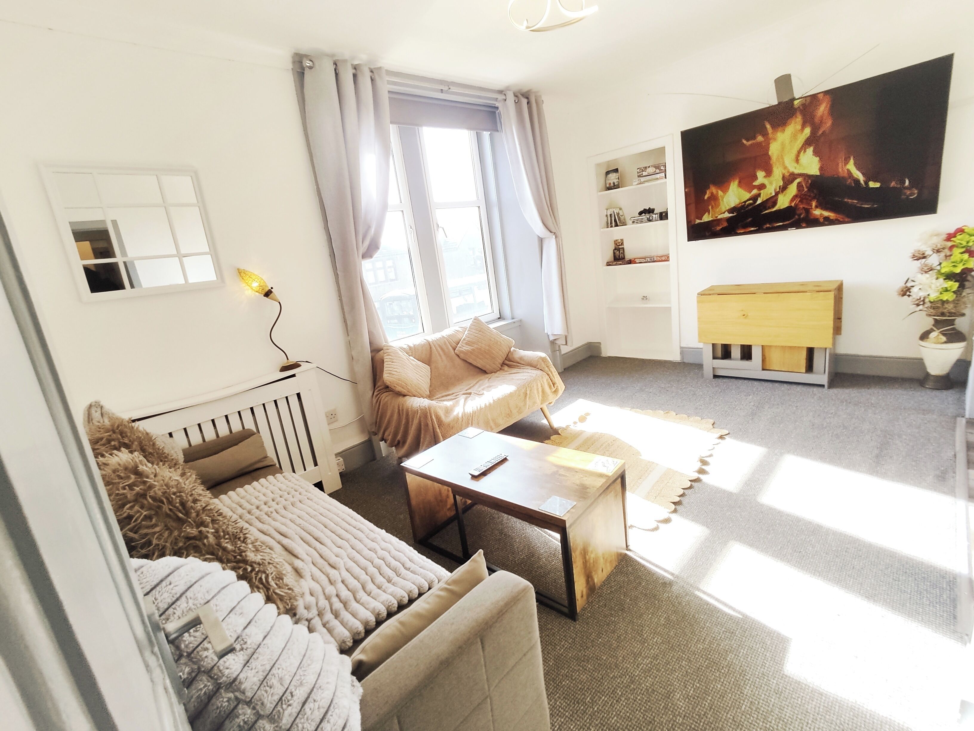 A living room with a sofa, a coffee table, a TV with a fire image, a radiator, and a window with curtains.