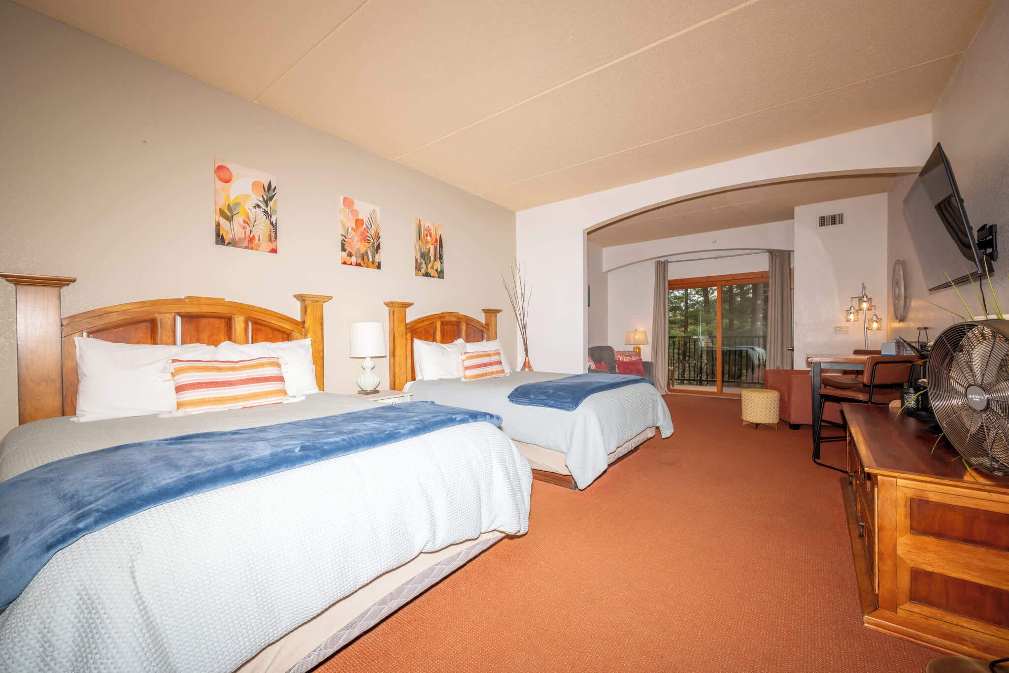 Habitación de hotel con dos camas, un escritorio, una silla, un televisor y obras de arte en las paredes.