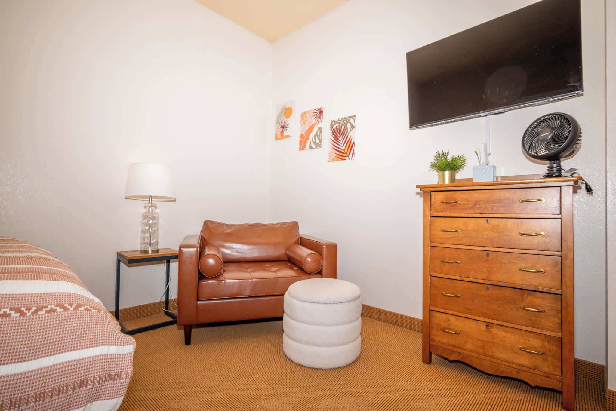 A bedroom with a brown leather sofa, a white ottoman, a wooden dresser, a TV mounted on the wall, and a small potted plant.