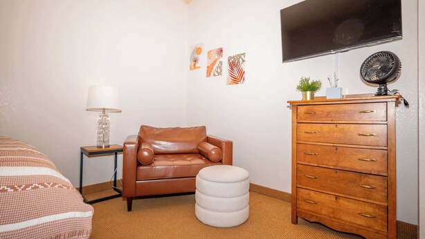 A bedroom with a brown leather sofa, a white ottoman, a wooden dresser, a TV mounted on the wall, and a small potted plant. - Chula Condo 2237 & 2239 (Wisconsin Dells)
