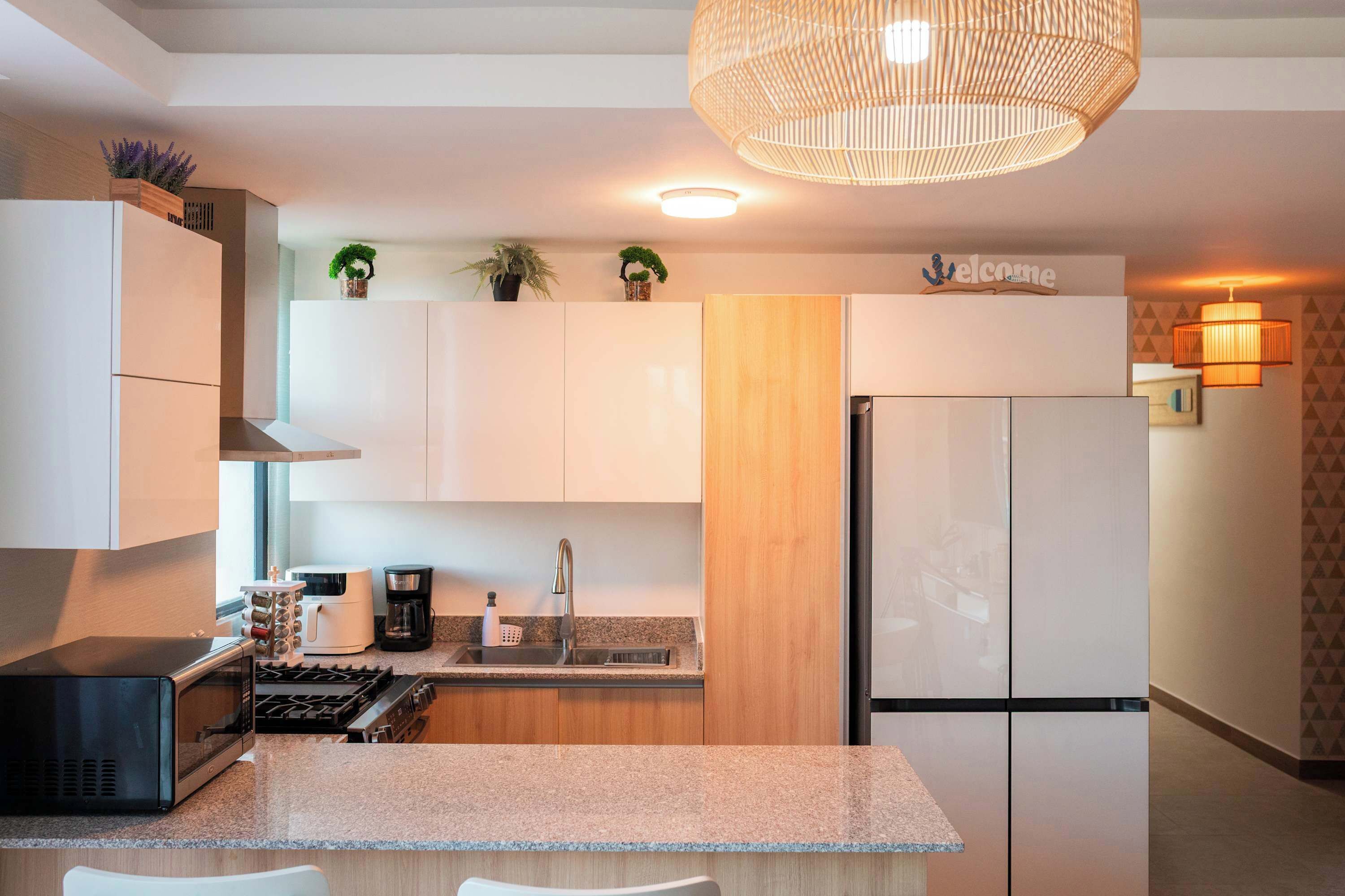 A modern kitchen with white cabinetry, a built-in stove, and a refrigerator. There are potted plants on the countertop and a hanging light fixture above the sink.