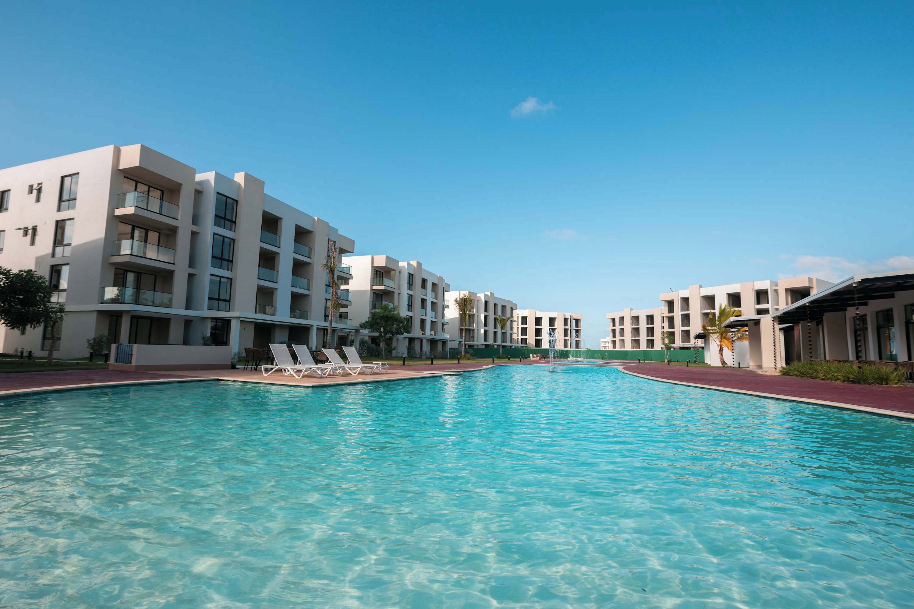 Een modern hotel met een centraal gelegen zwembad, meerdere balkons en een helderblauwe lucht.